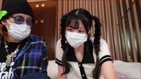 Media: A video of two women in face masks, one wearing glasses and a plaid shirt, the other with twin pigtails and a white blouse. They sit indoors, next to beige curtains.