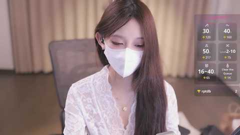 Media: Video of an Asian woman with long dark hair, wearing a white lace dress and face mask, sitting indoors with beige curtains and a black chair. A digital overlay shows a temperature of 15.4\u00b0C.