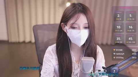 Media: Video of an East Asian woman with long brown hair, wearing a white mask, a light top, and white earphones, seated at a desk. Background features beige curtains and a partially visible microphone.