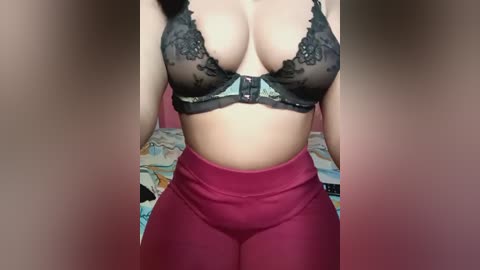 Media: Video of a woman with light skin, wearing a black lace bra and high-waisted, maroon leggings, standing in a bedroom with a bed and colorful, patterned bedding in the background.