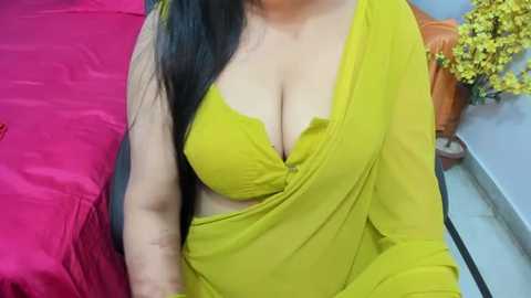 Media: A video of a woman with long black hair, wearing a bright yellow saree that reveals a lacy green bra, seated on a bed with a red blanket and a potted plant in the background.