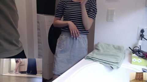 Media: A video of a woman in a striped shirt and light gray skirt, holding a phone, taking a mirror selfie in a modern bathroom.