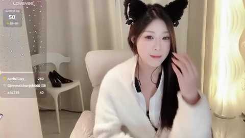 Media: Video of an Asian woman with long black hair, wearing black cat ears and a white robe, sitting in a modern room with white furniture and a lamp.