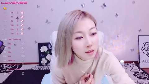 Media: A video of an Asian woman with light skin and straight, shoulder-length blonde hair, wearing a beige turtleneck sweater. She's sitting in front of a white wall with butterfly stickers. The background includes a calendar and a black floral design.