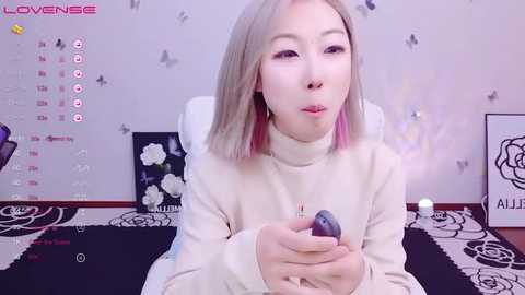 Media: Video of an Asian woman with straight, shoulder-length platinum blonde hair, wearing a cream-colored turtleneck sweater, holding a smartphone. Background features a white wall with black floral and butterfly decals, a calendar, and a black and white rug.