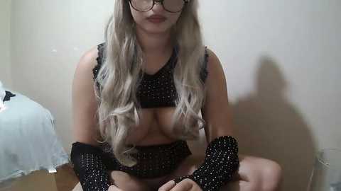 Media: A video of a light-skinned woman with long, wavy blonde hair, wearing black polka-dotted lingerie, sitting cross-legged on a wooden floor in a dimly lit room. She has large breasts and wears glasses.