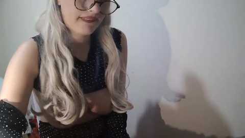 Media: Video of a smiling young woman with long, wavy platinum blonde hair, wearing glasses and a black polka-dotted crop top, standing against a plain white wall.