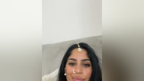 Media: A video of a smiling woman with long black hair, wearing a gold forehead ornament, a nose ring, and white earrings, against a plain white wall.