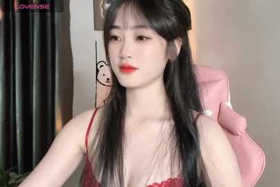 Media: Video of an East Asian woman with fair skin, long black hair, and red lipstick, wearing a red lace bra, seated in a pink chair in a modern room.