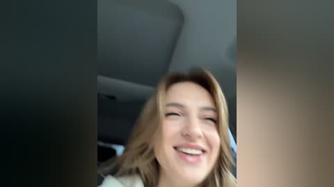 Media: A video of a smiling, fair-skinned woman with wavy blonde hair, wearing a white top, seated in a car with a grey headrest visible in the background.