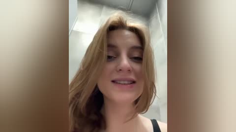 Media: Video of a young Caucasian woman with light skin and shoulder-length, wavy blonde hair, smiling, wearing a black top, standing in a modern bathroom with white tiled walls and a ceiling.