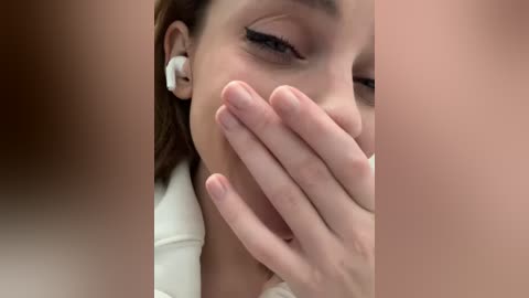 Media: A close-up video of a young woman with light skin, wearing a white top and white earbuds, covering her mouth with her hand, eyes closed, suggesting she is coughing or sneezing.