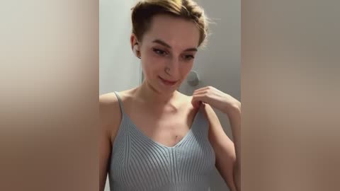 Media: Video of a young woman with fair skin, light brown hair tied back, wearing a light gray ribbed tank top, smiling subtly, and standing in front of a blurred beige background.