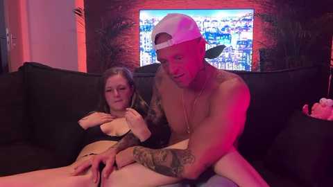 Media: A video shows a tattooed, shirtless man with a backward cap caressing a nude woman with large breasts on a black couch, with a TV displaying a sports game in the background.