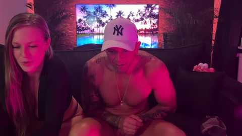 Media: Video of a shirtless, tattooed man wearing a New York Yankees cap, sitting on a couch with a blonde woman, both looking relaxed in a dimly lit room with a tropical beach scene TV background.