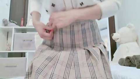 Media: Video of a person in a plaid skirt, adjusting their waistband, in a minimalist, white-walled room with a bed, teddy bear, and storage shelves.