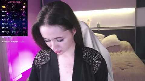 Media: A video of a fair-skinned woman with shoulder-length black hair, wearing a black lace robe, standing in a dimly lit bedroom with a bed and nightstand visible in the background.