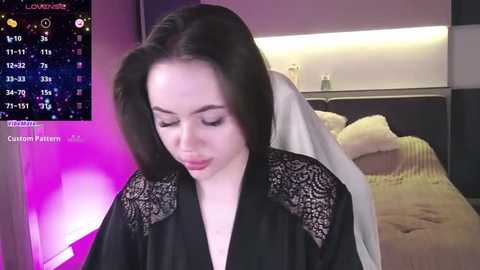 Media: A video of a fair-skinned woman with long black hair, wearing a black lace robe, standing in a modern bedroom with a bed and digital calendar in the background.