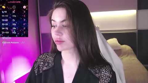 Media: Video of a young woman with straight, shoulder-length brown hair in a black lace-trimmed robe, standing indoors with a purple light in the background.