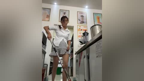 Media: A video of a woman in a white blouse and checkered skirt cleaning a kitchen with stainless steel appliances and colorful posters on the wall.