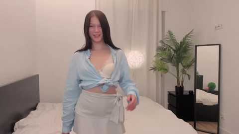 Media: Video of a smiling woman with long brown hair, wearing a blue shirt tied at the waist, and a white apron, standing in a minimalist bedroom with white walls, a bed, and a green potted plant.