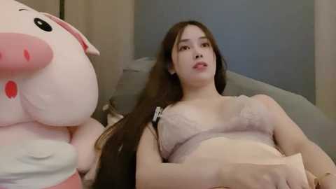 Media: Video of an East Asian woman with long black hair, wearing a light pink lace bra, lying on a bed with a plush pig toy.