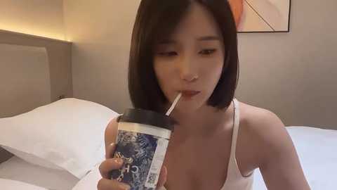 Media: Video of a young East Asian woman with straight black hair, drinking from a blue and white coffee cup in a minimalist bedroom.