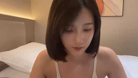 Media: A video of a young Asian woman with short black hair, wearing a white tank top, sitting on a bed with white linens. The background shows a beige wall and an abstract painting.