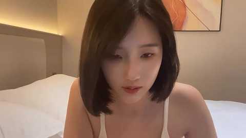 Media: Video of an East Asian woman with shoulder-length black hair, fair skin, and red lipstick, wearing a white tank top, sitting on a bed in a modern, beige-walled room with minimalistic decor.