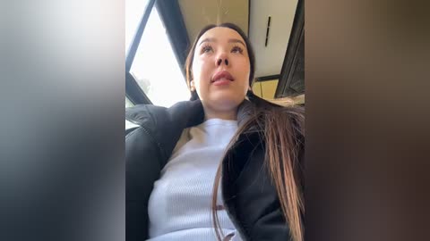 Media: Video of a young woman with light skin, long dark hair, and a white top, seated in a car, looking upward.