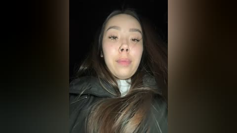 Media: Video of a young Asian woman with long brown hair, wearing a black jacket, making a pouty face against a dark background.