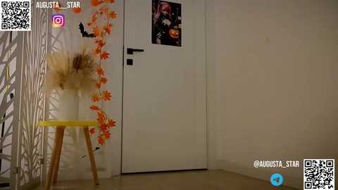 Media: Video of a minimalist room with a white door, a yellow stool, and a tall vase of dried flowers on the left. The background features an orange leaf decoration and a small framed picture. QR codes and a watermark are visible.