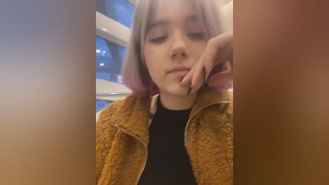 Media: A video of a young woman with light skin and shoulder-length pastel purple hair, wearing a mustard-yellow fleece jacket over a black top. She has a contemplative expression, with her hand resting near her mouth. The background shows a blurred blue and white interior.