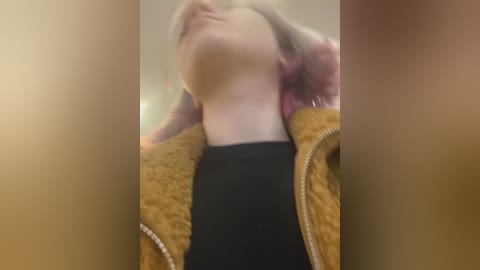 Media: Video of a person with light skin, wearing a black shirt and a brown, fuzzy jacket, seen from the neck up, slightly blurred background.