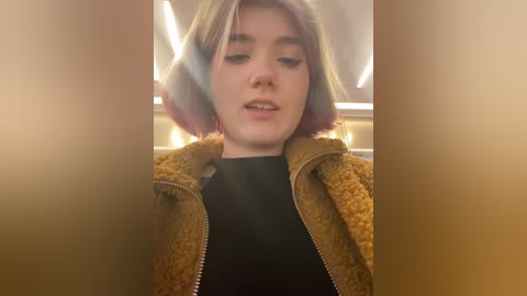 Media: Video of a young woman with shoulder-length, light blonde hair and fair skin, wearing a mustard-yellow, textured jacket over a black top. Background is blurred, showing an indoor setting with warm lighting.