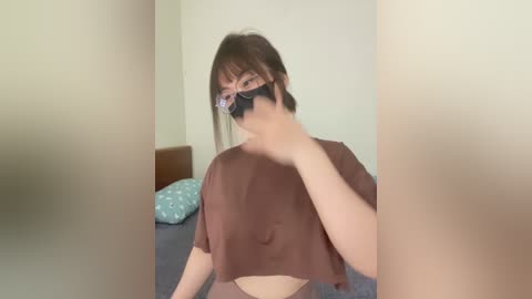 Media: A video of a young woman with short brown hair, wearing a brown crop top and black face mask, standing in a minimalistic bedroom with a beige wall and a bed with a blue pillow.