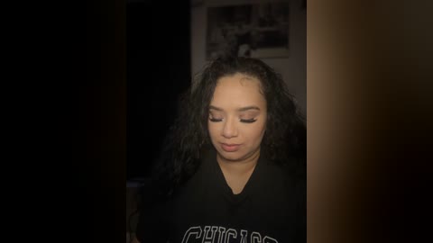 Media: Video of a young woman with curly black hair, wearing a black t-shirt, looking down thoughtfully in a dimly lit room with a framed picture on the wall in the background.