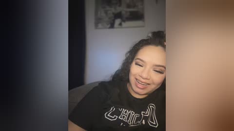 Media: Video of a woman with curly hair, smiling, wearing a black t-shirt with white text, leaning against a dark wall. Background features a framed black-and-white video.