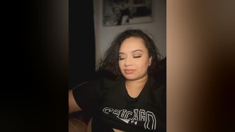 Media: Video of a Latina woman with curly black hair, light skin tone, and medium build, wearing a black t-shirt, sitting indoors, eyes closed, serene expression.