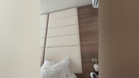 Media: A video of a minimalist bedroom with a wooden headboard, white pillows, and a beige wall. The room is modern and clean, with a hint of a white bedside lamp.