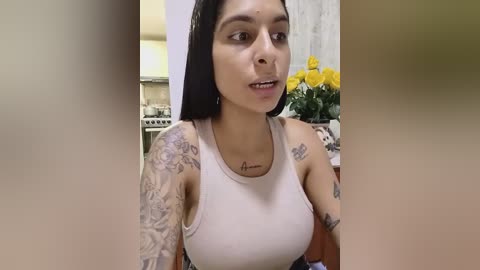 Media: A video of a Latina woman with medium brown skin, long black hair, and tattoos, wearing a white tank top, in a kitchen with yellow flowers and a stove in the background.