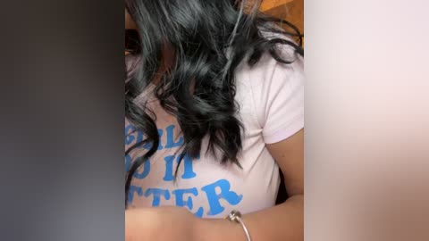 Media: Video of a woman with wavy black hair, wearing a white T-shirt with blue text partially covering her breasts. Her face is not visible, and she's holding a silver bracelet.