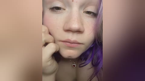 Media: Video of a young woman with pale skin, light freckles, and shoulder-length purple hair. She has a neutral expression, resting her chin on her hand. The background is blurred and pinkish.