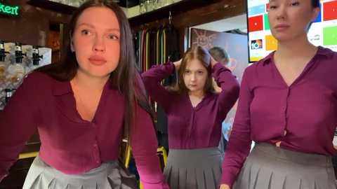 Media: Video of three women in a clothing store, wearing matching maroon cardigans and grey pleated skirts, adjusting their hair. Background includes colorful posters and clothing racks.