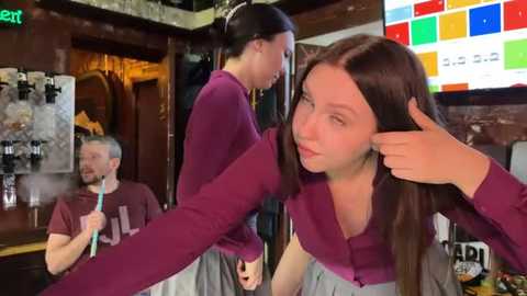 Media: Video of a young woman with long brown hair, wearing a maroon sweater and grey pants, playfully pulling her hair while another woman in maroon stands beside her, both in a cozy, dimly-lit pub setting.