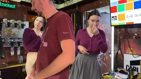 Media: A video of a young woman with long dark hair in a burgundy top and grey skirt covering her mouth in a bar with a mirror reflecting her, a man in a burgundy shirt and glasses, and a colorful TV screen.