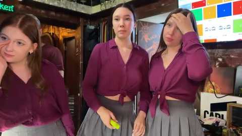 Media: Video of three young women with long hair, wearing maroon tops tied at the waist and grey pleated skirts, standing in a dimly lit bar, one adjusting her hair.