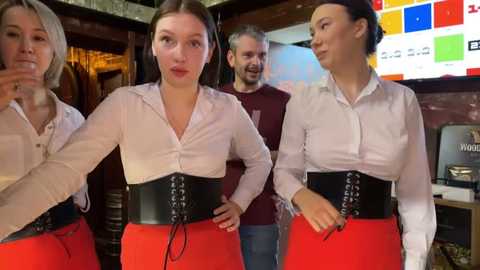 Media: Video of four young women in white blouses, red skirts, and black corsets, posing in a dimly lit, cozy bar. Background includes a man and colorful, abstract artwork.