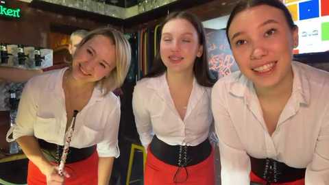 Media: Video of three smiling young women, one blonde, one brunette, and one with brown hair, wearing white blouses and red skirts, in a dimly-lit bar with a \"kert\" sign, beer taps, and a colorful background.