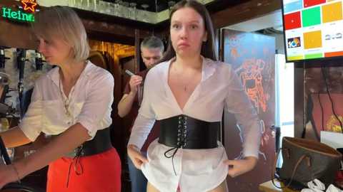 Media: Video of two women in a bar: one in a white blouse with a black corset, the other in a red dress. Background shows a neon \"Heineken\" sign, bar decor, and a TV screen.